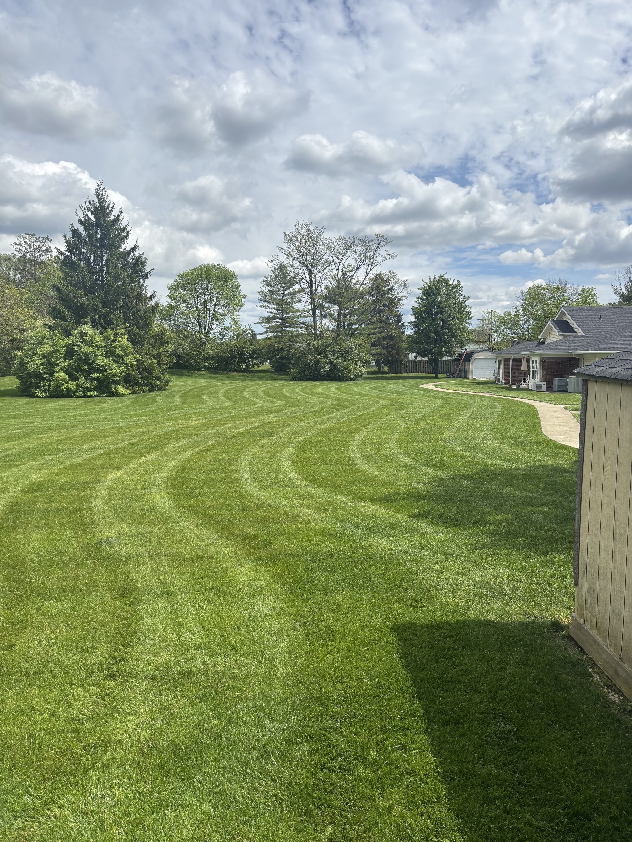 Well-maintained green lawn with visible mowing lines, trees, and a house in the background under a partly cloudy sky.