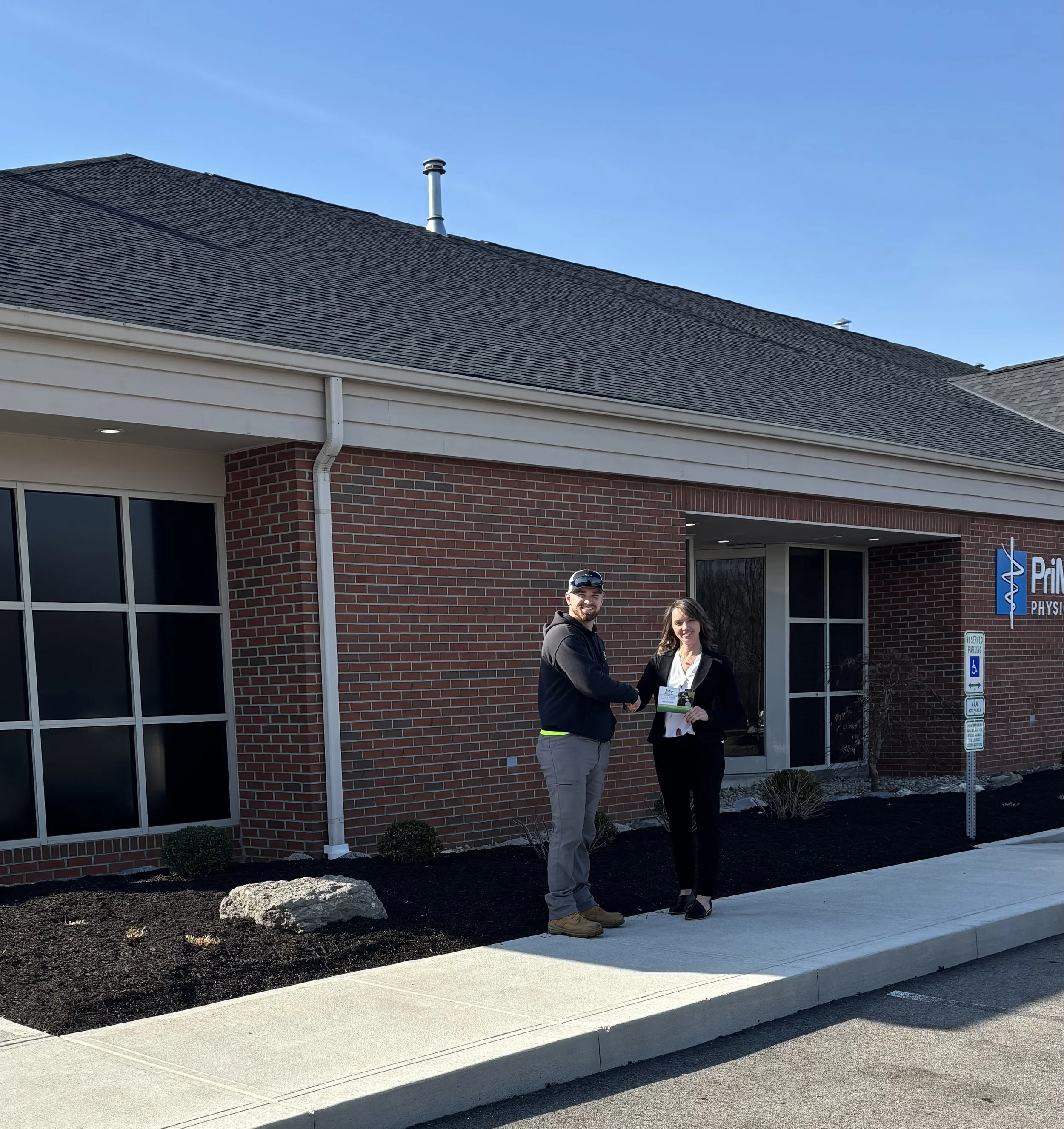 A man and a woman shaking hands outside a brick building, with the woman holding a folder, under a sign that reads 'Prime Physical Therapy'.