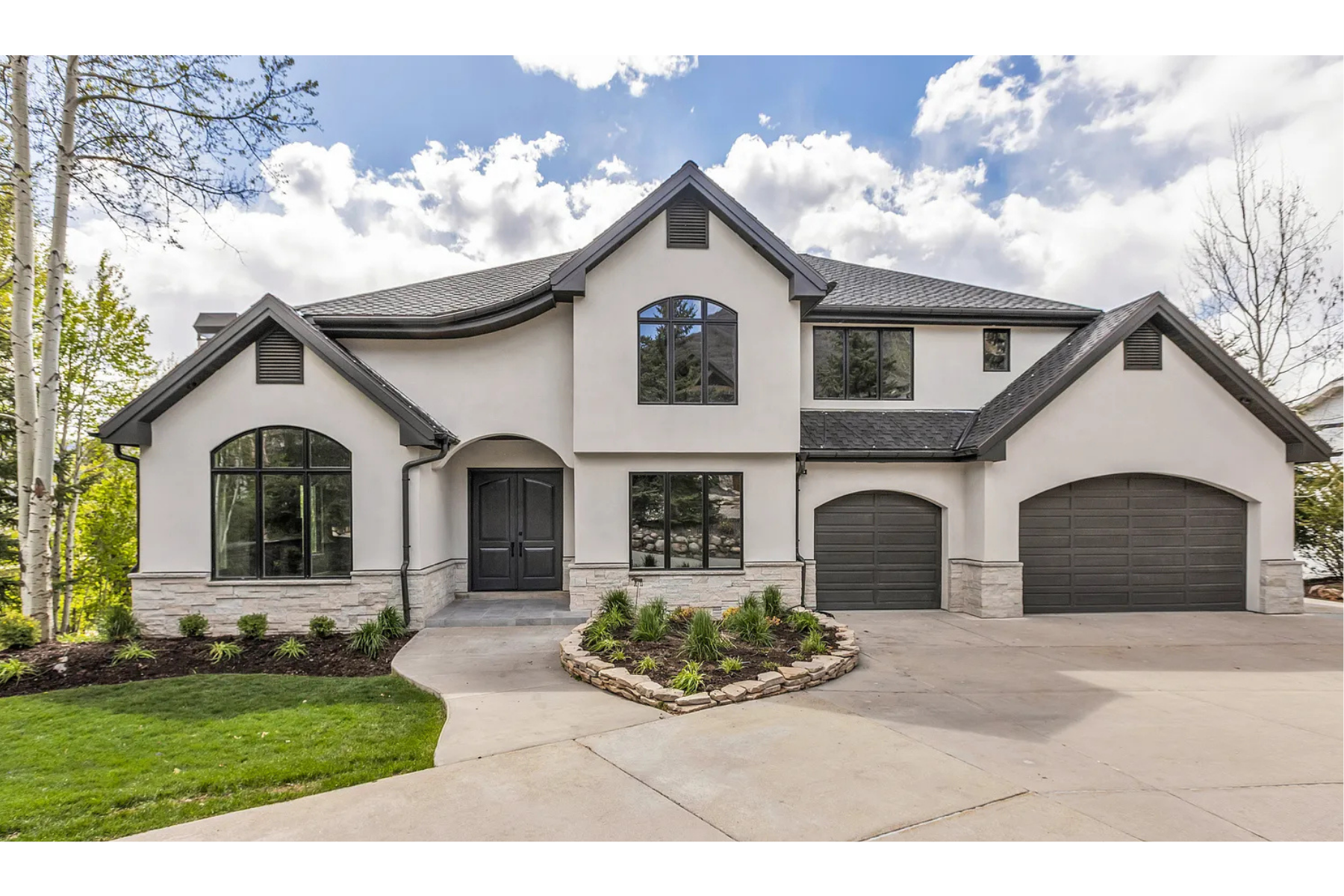 Modern two-story house with white stucco exterior, black front door, large windows, and a dark gray roof. Manicured lawn and landscaped front yard with small plants. Attached three-car garage and driveway.