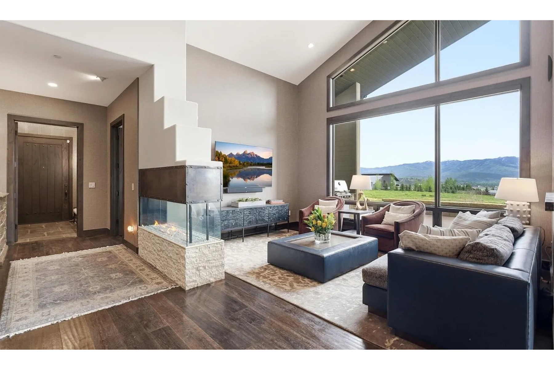 Bright living room with large windows showing mountains and green landscape. Contains a black leather sofa with throw pillows, two plush armchairs, a coffee table with a flower arrangement, a modern fireplace, wall-mounted TV, and side lamps. Hardwood floors and area rugs complete the cozy decor.