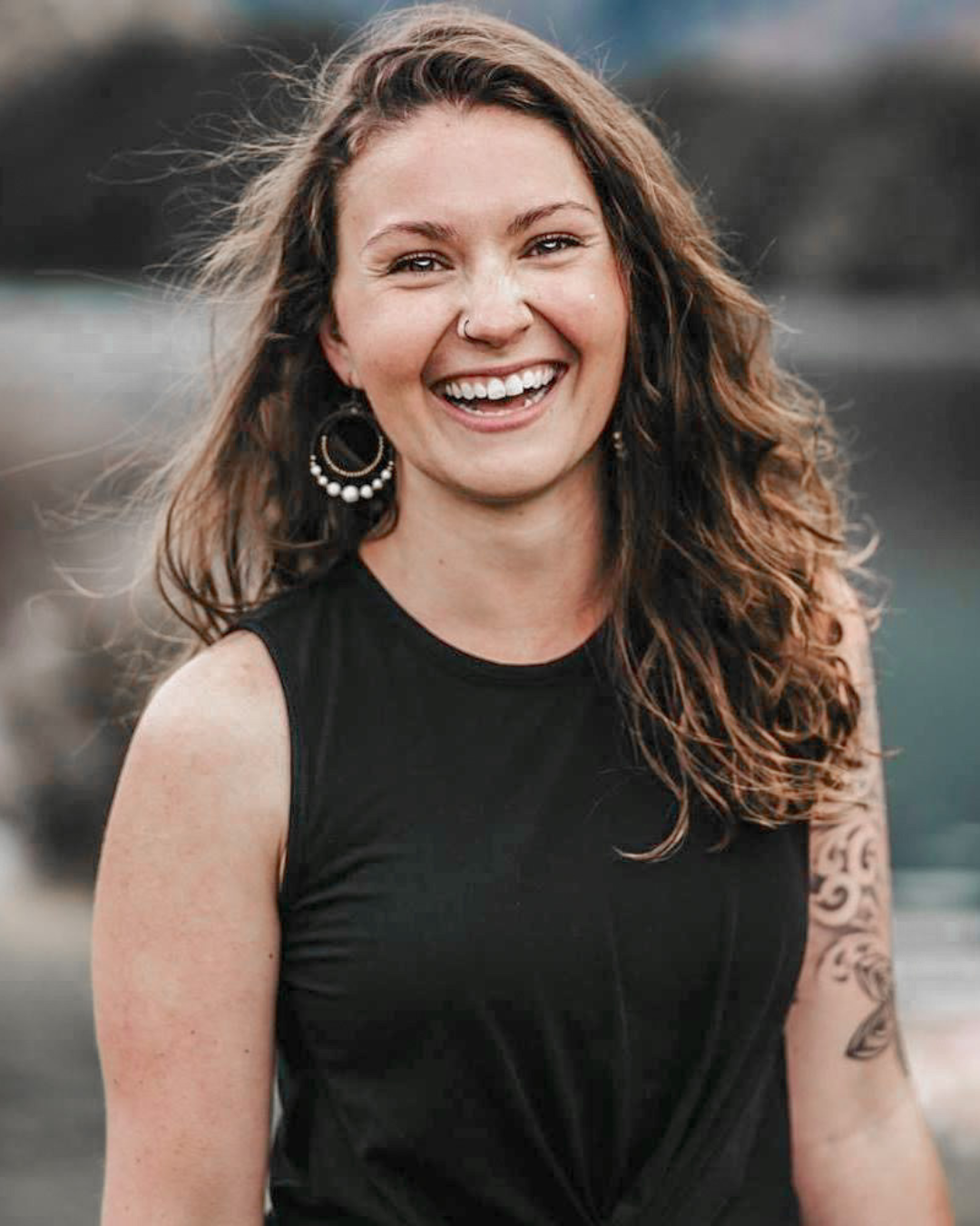 A young woman with curly brown hair, wearing a black sleeveless top, smiling, and wearing earrings with a hoop and pearl design, outdoors with a blurry background.