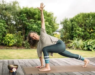 A woman practicing yoga in a seated lotus pose, smiling, wearing a white t-shirt and a blue jacket indoors with a concrete wall background.