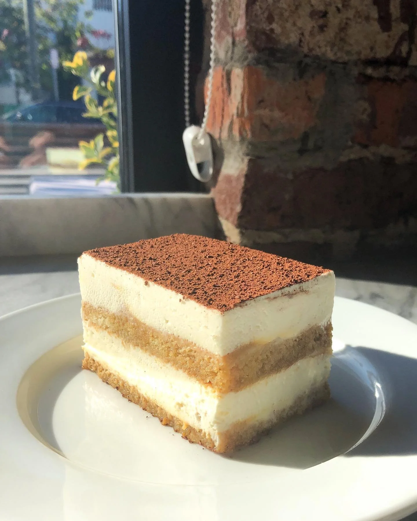 Slice of tiramisu on a white plate near a window with brick wall backdrop.