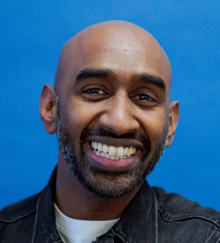 Smiling man with a beard, bald head, wearing a denim jacket against a blue background.