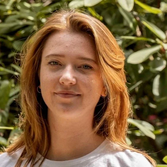 Person with red hair in an outdoor setting with green leaves in the background.