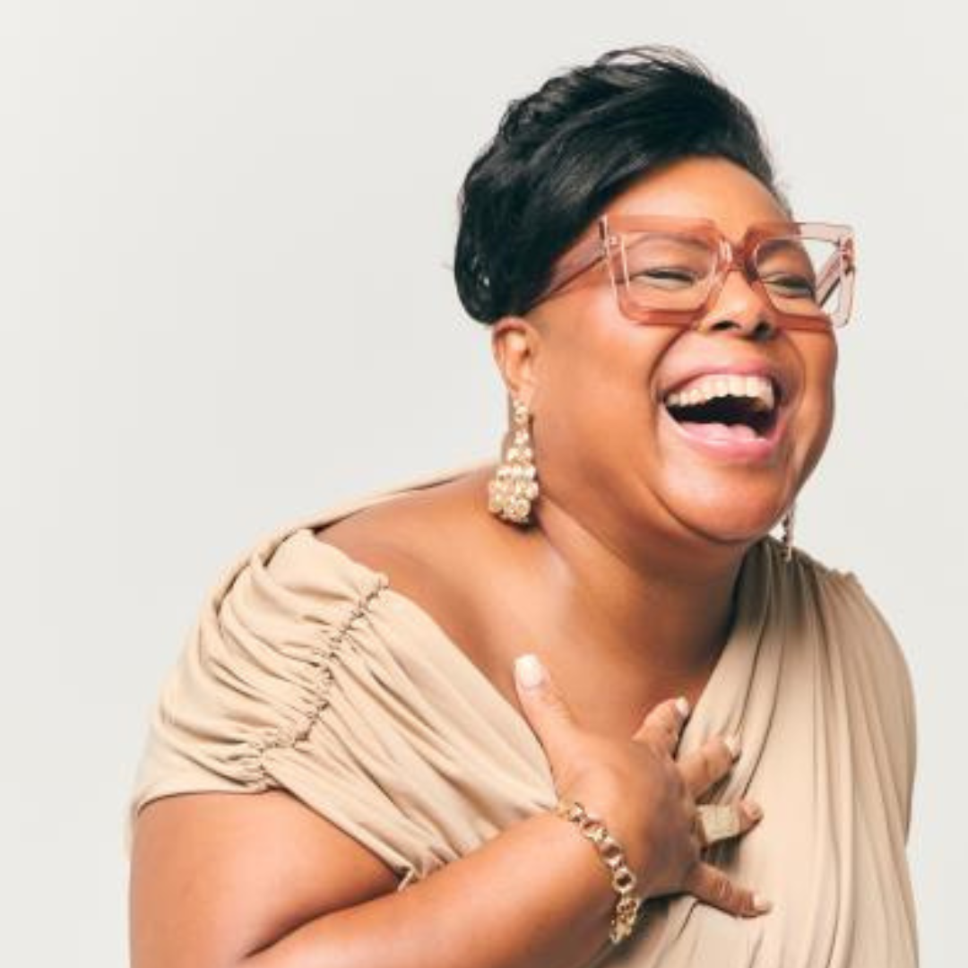 Person laughing joyfully, wearing glasses and a beige dress with jewelry.