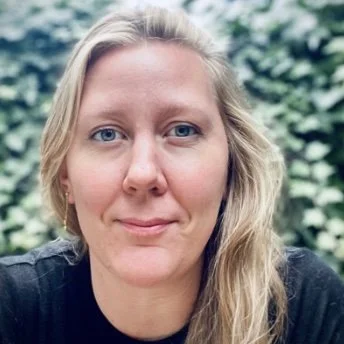 Person with long blonde hair and blue eyes, wearing a black shirt, with a leafy background.