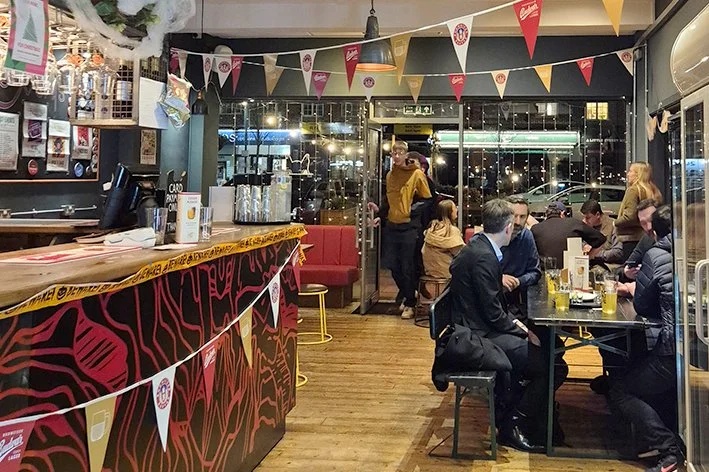 The image shows a lively bar or restaurant with customers dining and socializing. The space is decorated with colorful bunting hanging from the ceiling, and large windows offer a view of the street outside. There is a bar counter on the left and seve