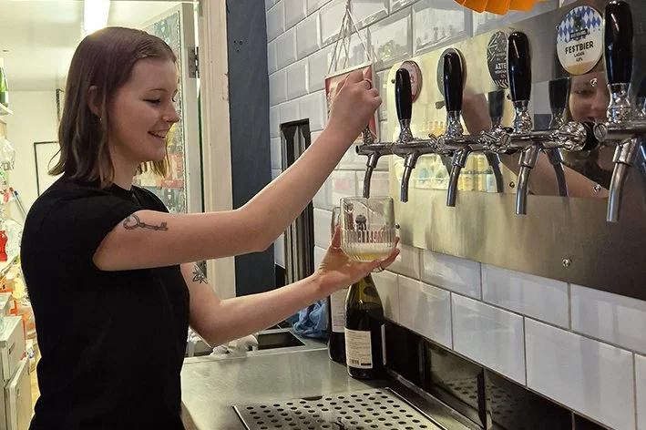 A woman with shoulder-length brown hair pouring beer from a tap into a glass at a bar or brewery, smiling, with a mirror reflecting her face in the background.