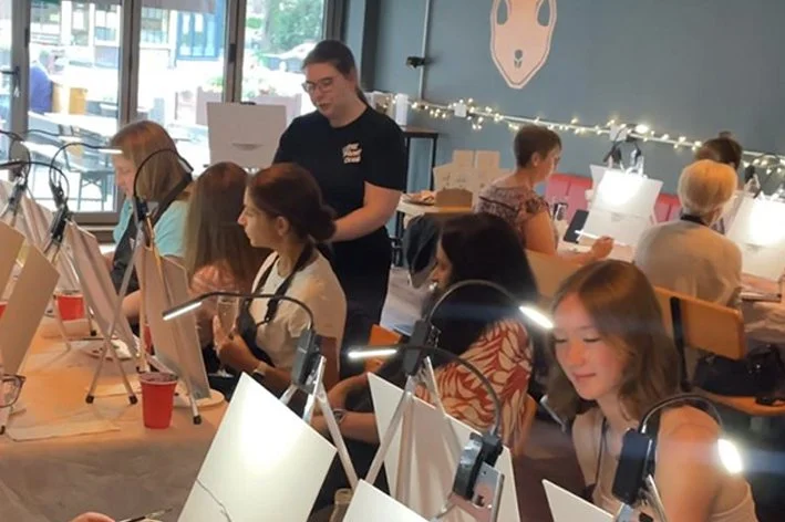 Group of women painting at a social art class with an instructor guiding them, in a decorated room with string lights.