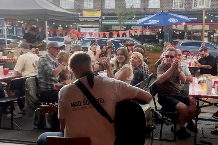 A man with a guitar performing for a crowd at an outdoor restaurant or bar during the daytime.