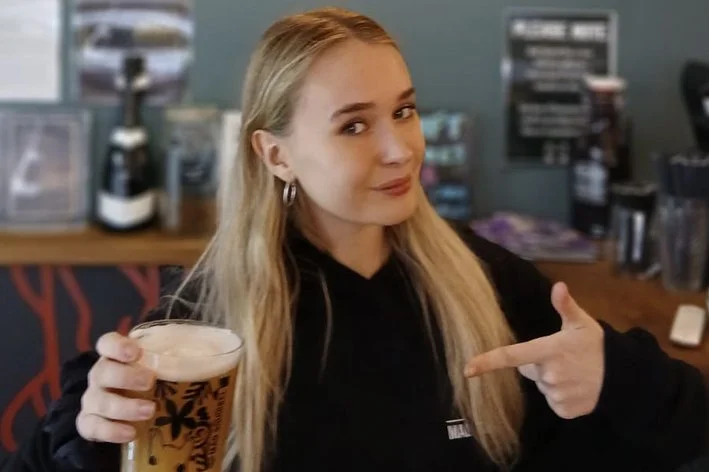 A young woman with long blonde hair and hoop earrings points at a glass of beer in a bar or pub setting.