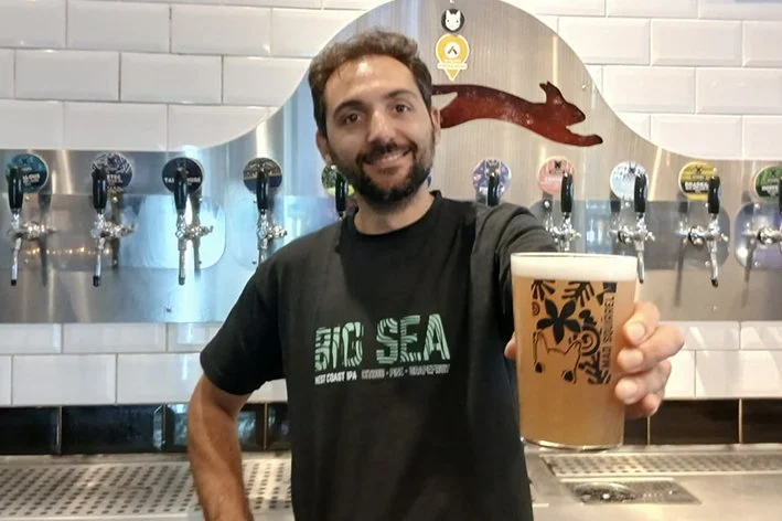 Man smiling and holding a glass of beer in front of a row of beer taps at a bar.