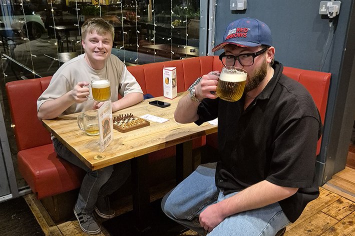 Two men sitting at a wooden table in a restaurant, each holding a glass of beer. The man on the left is smiling, wearing a beige T-shirt, and the man on the right is wearing glasses, a baseball cap, and a black shirt. They are mid-meal. The table has