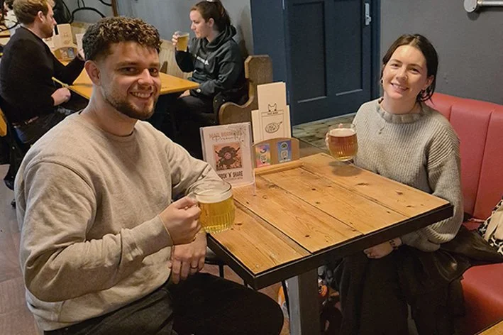 A man and woman sitting at a wooden table in a restaurant, each holding a glass of beer, smiling at the camera.