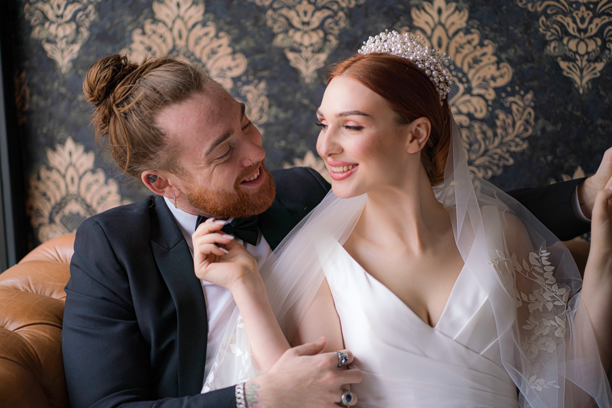 Bride and groom smiling at each other on a couch with floral wallpaper background.
