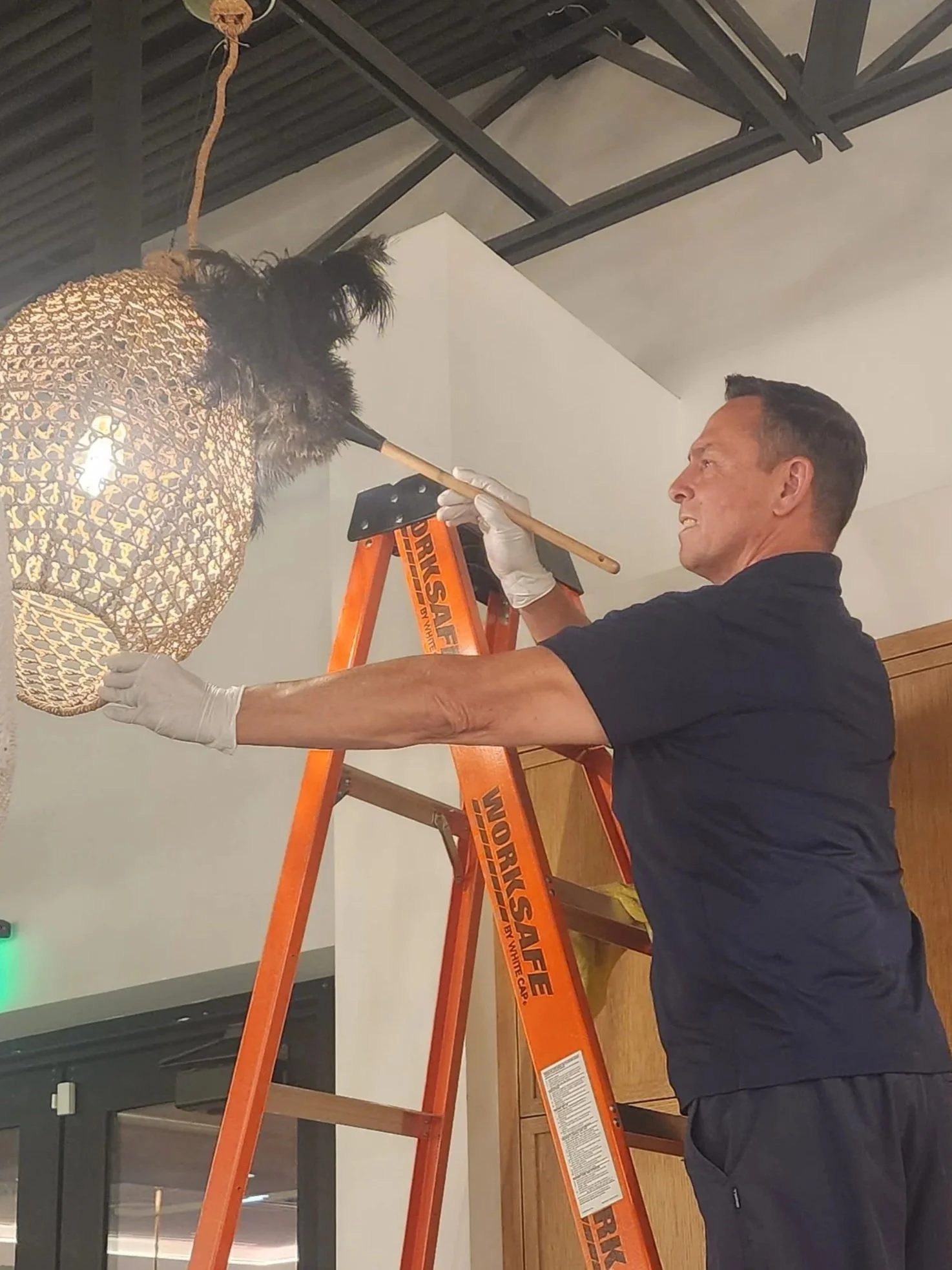 A man with short dark hair wearing a black shirt and white gloves on both hands, standing on an orange ladder, is cleaning or inspecting a decorative wicker pendant light with black feather accents, suspended from the ceiling.