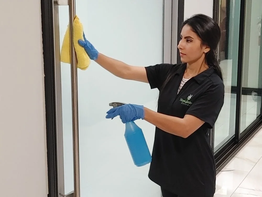 A woman in a black uniform and blue cleaning gloves using a yellow cloth to clean glass door, holding a blue spray bottle.