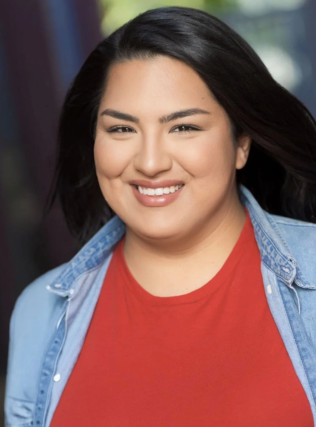 A woman with long black hair, brown eyes, wearing an orange shirt and blue denim shirt, looking at the camera with a slight smile.