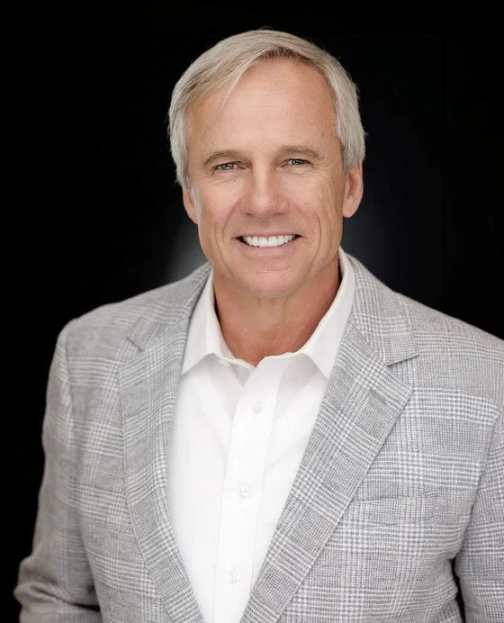A man with white hair and blue eyes, wearing white shirt and gray coat, standing against a black background.