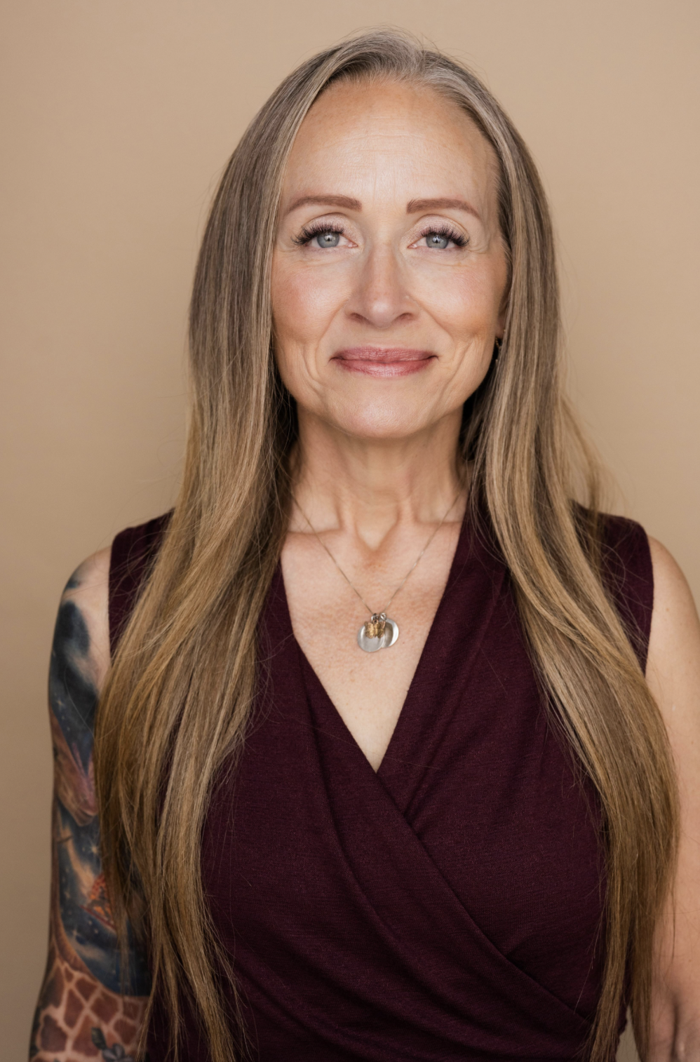 A woman smiling with long hair and a plum blouse, against a tan background.