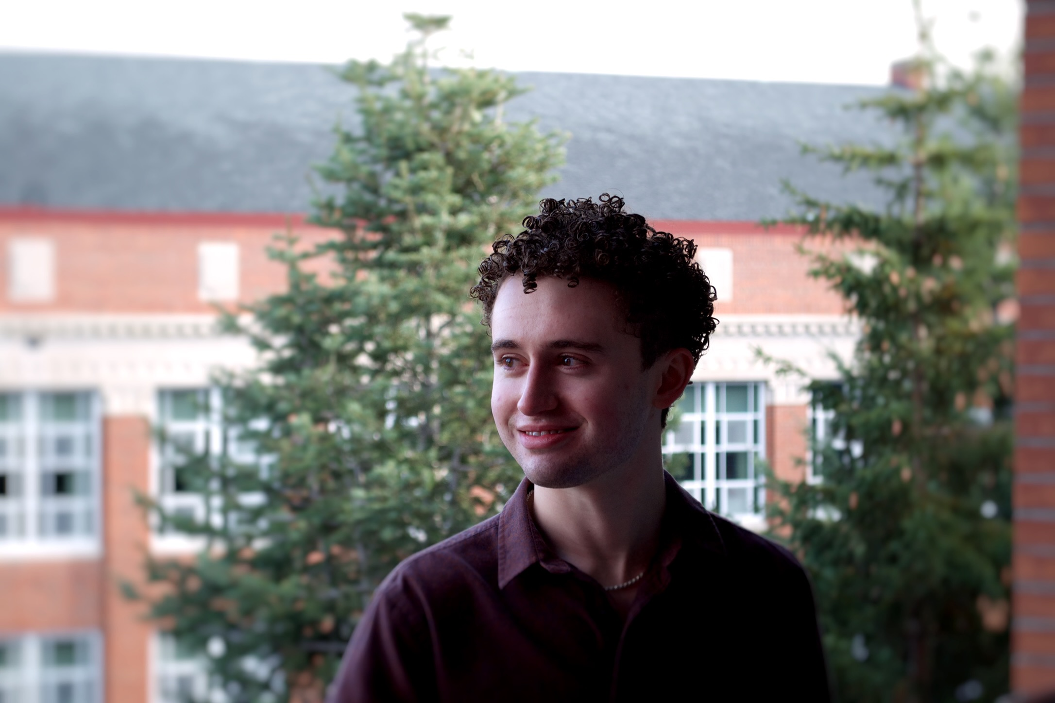 A young man with curly dark hair wearing a dark shirt, standing outdoors with trees and a red-brick building in the background.