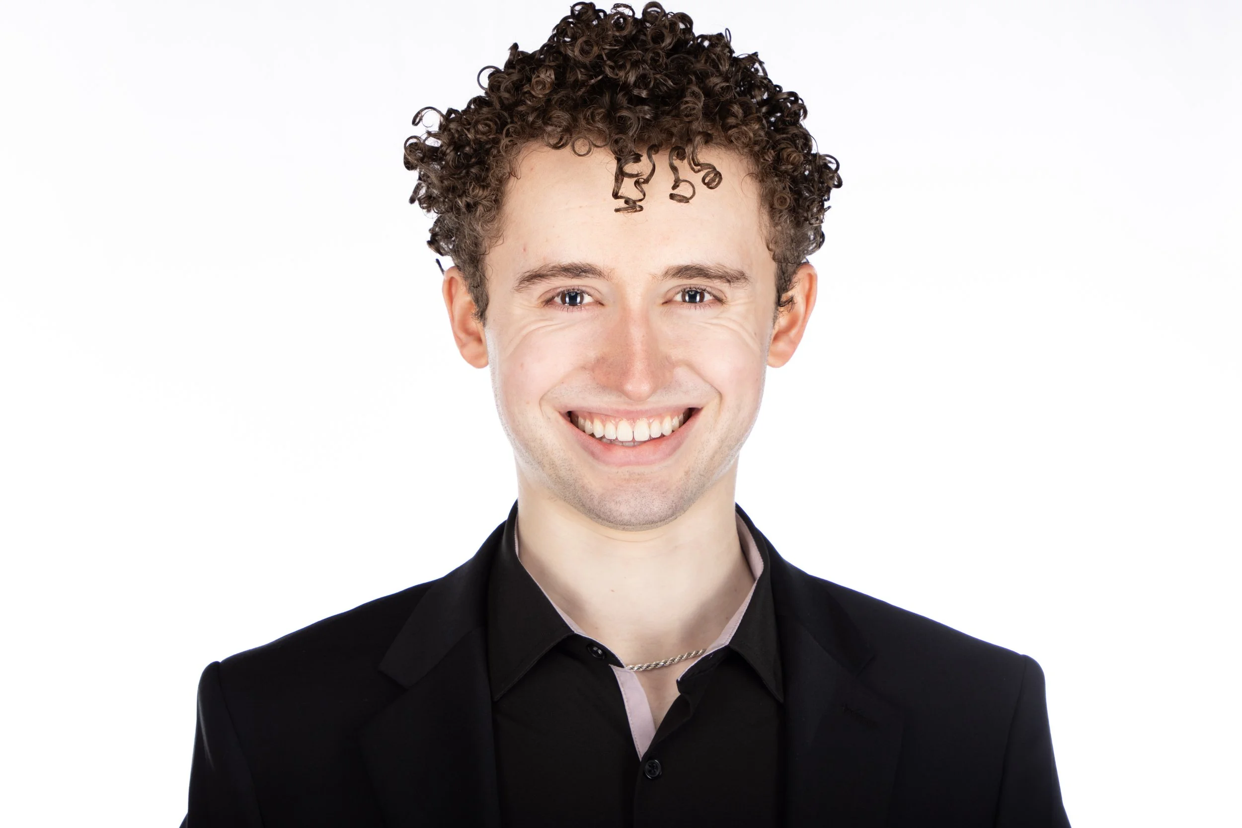 Headshot of a young man with curly brown hair, smiling, wearing a black suit jacket and a black shirt, against a white background.