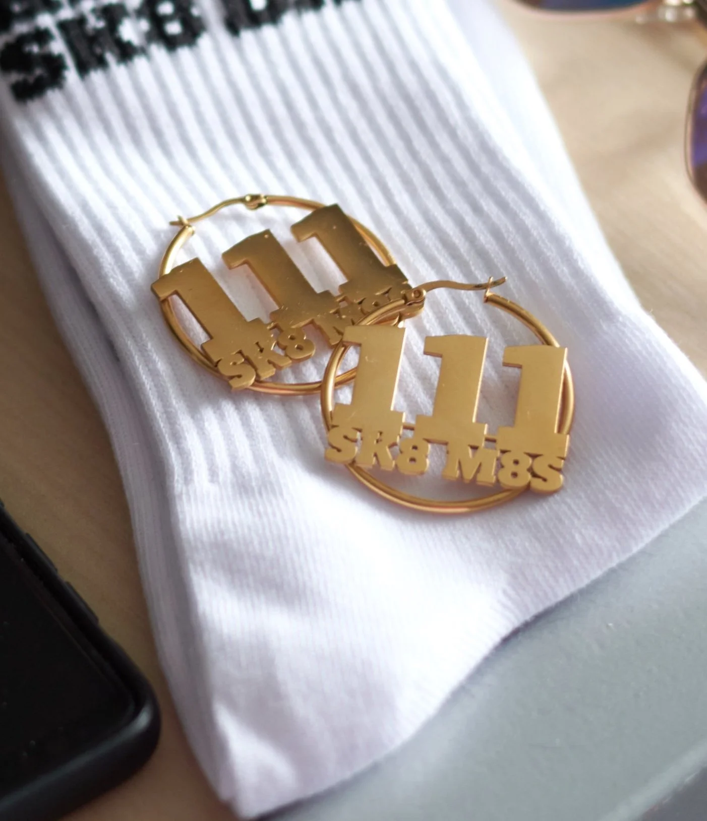 Detailed view of SK8 M8S Gold hoop earrings with '111' and 'SK8 M8S' letter cutouts, placed on white folded SK8 M8S roller skate socks with black text