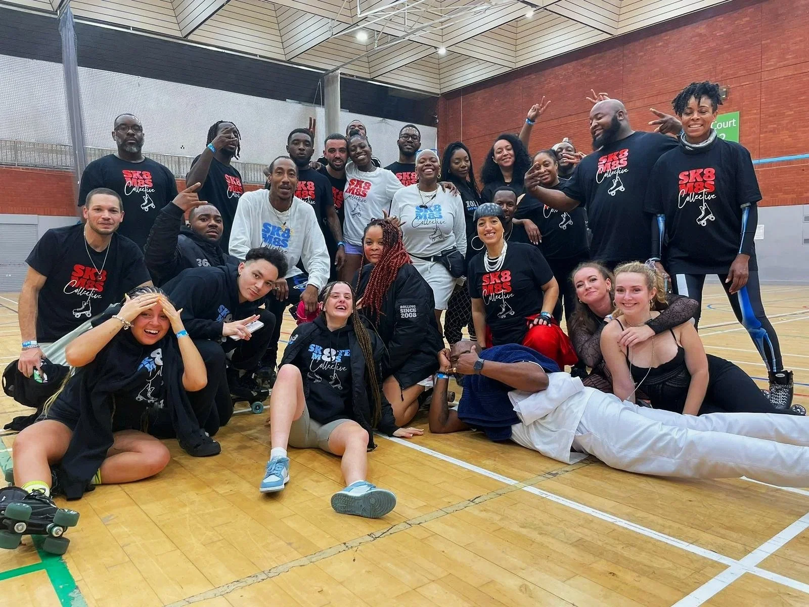 A large group of people roller skaters, diverse in age and ethnicity, gathered in a gymnasium, posing and smiling at the camera. Some are standing, others are sitting or lying on the floor, and many are wearing matching sk8 m8s classic t-shirts