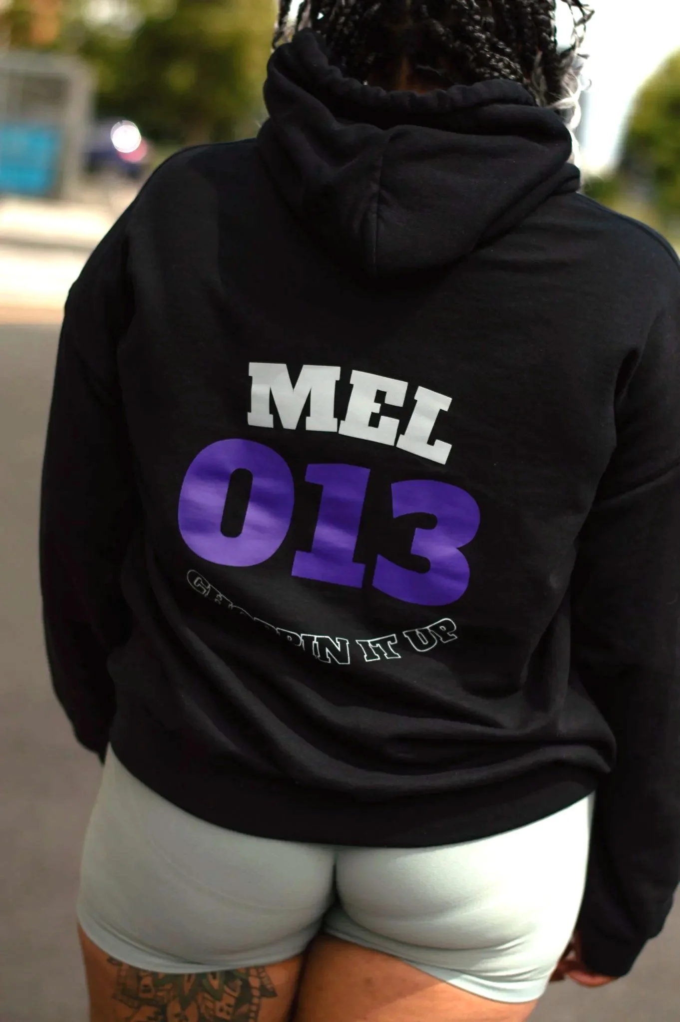 Roller skater back to camera wearing black SK8 M8S hoodie with purple SK8 No. 013 and light grey reflective text Mel, light green shorts, standing outdoors.