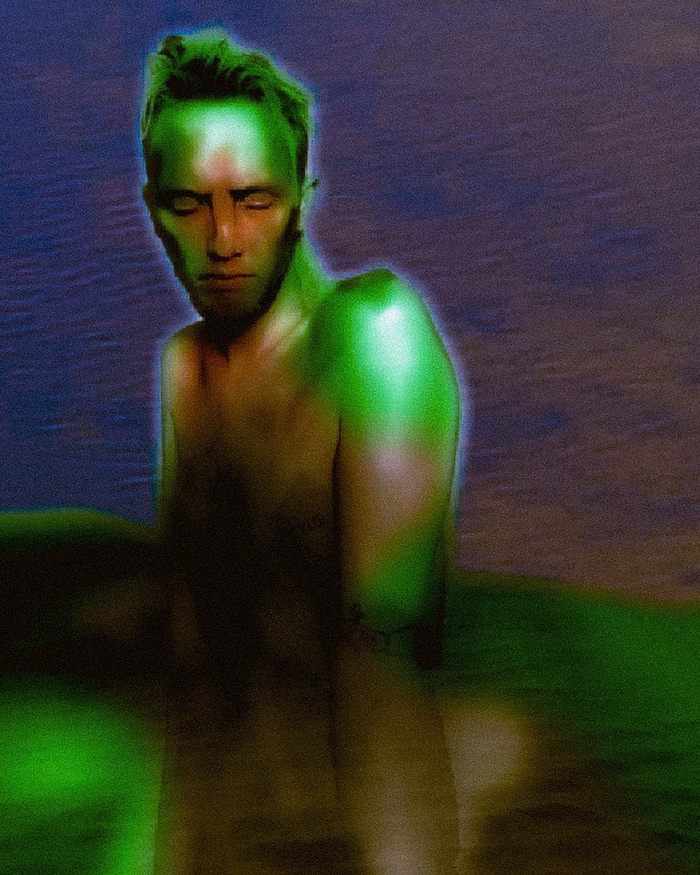 Photo of a shirtless man with light skin and styled hair posing against a water background, with green digital effects on his shoulders and arms.