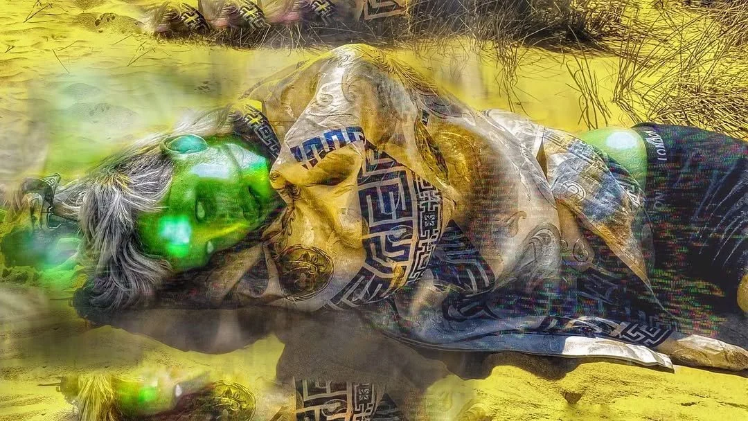 A person with gray hair lying on the ground with eyes closed, wearing sunglasses, a patterned shirt, and a scarf, in a natural outdoor setting with yellowish grass and plants.