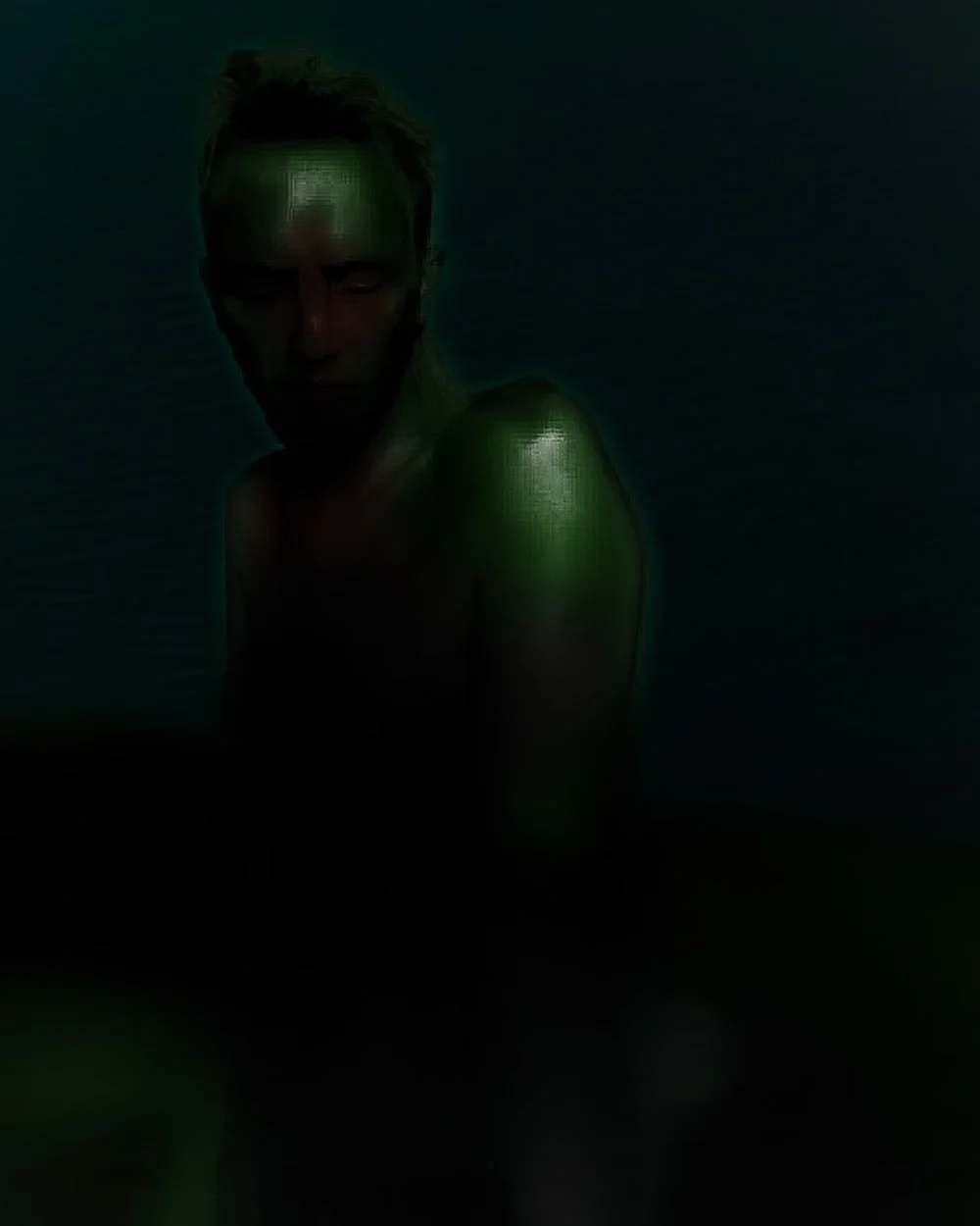 A person with short hair illuminated with green light, sitting in a dark environment with a serious expression.