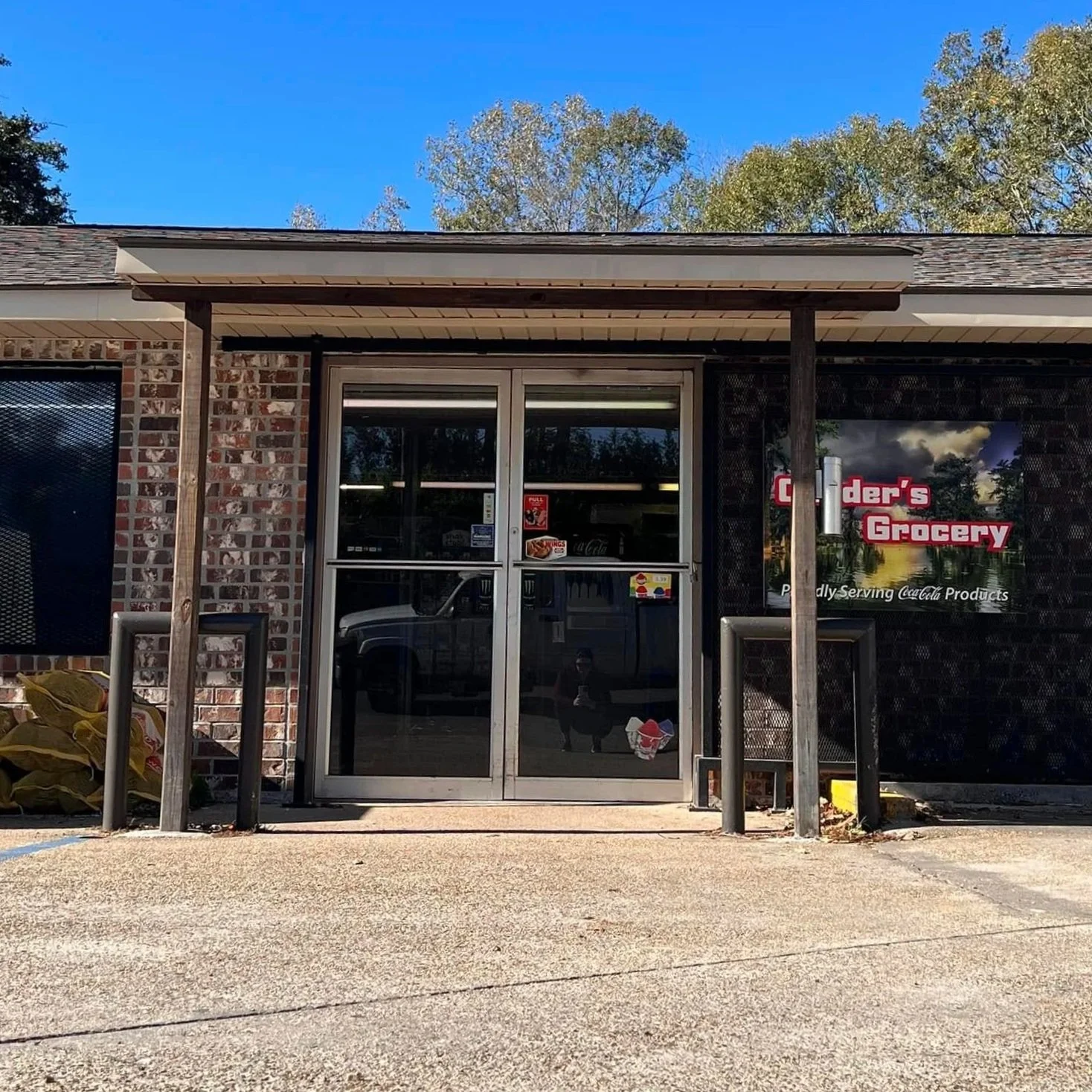 Front view of a small grocery store called 'Cinder's Grocery' with brick and black exterior walls, glass double doors, and a small parking area in front. The store has a sign with Coca-Cola branding and a few stickers on the door. Trees and blue sky are visible in the background.