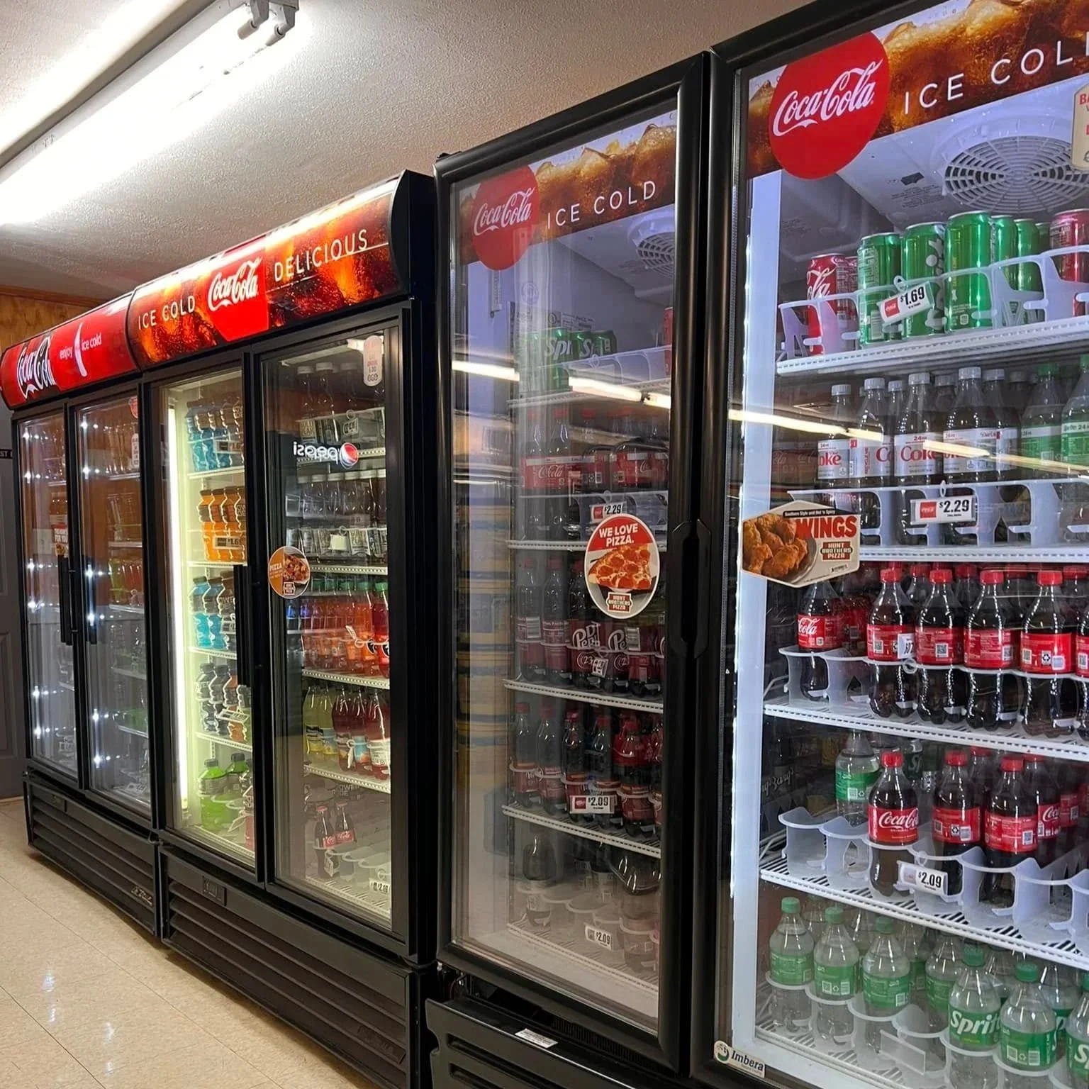 A row of commercial refrigerators in a convenience store, stocked with soda bottles and cans, including Coca-Cola, Sprite, and Diet Coke.