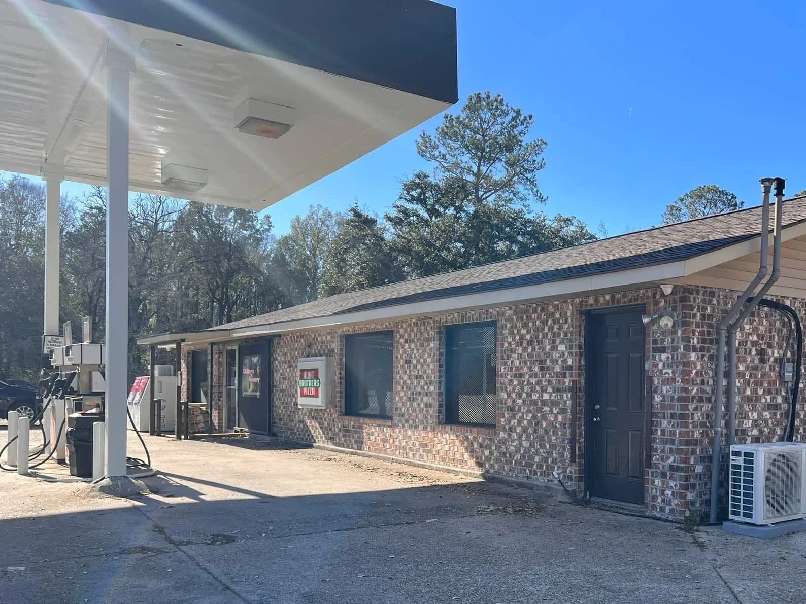 Gas station with multiple fuel pumps under a canopy, positioned next to a brick building with a sign advertising pizza, surrounded by trees and a clear blue sky.