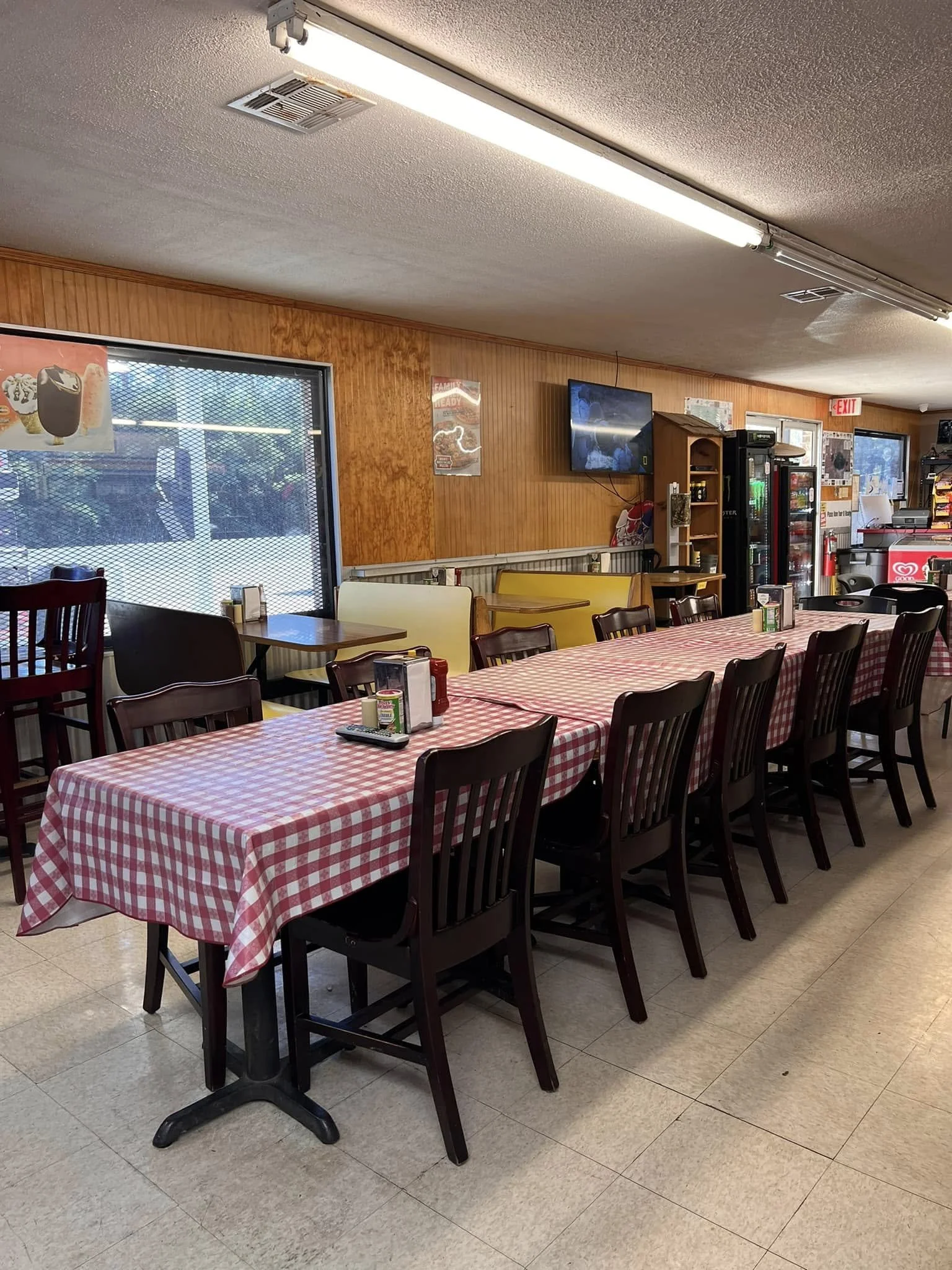 Interior of a casual restaurant with a long table covered with a red and white checkered tablecloth, surrounded by chairs, with condiments and a TV on the wall.