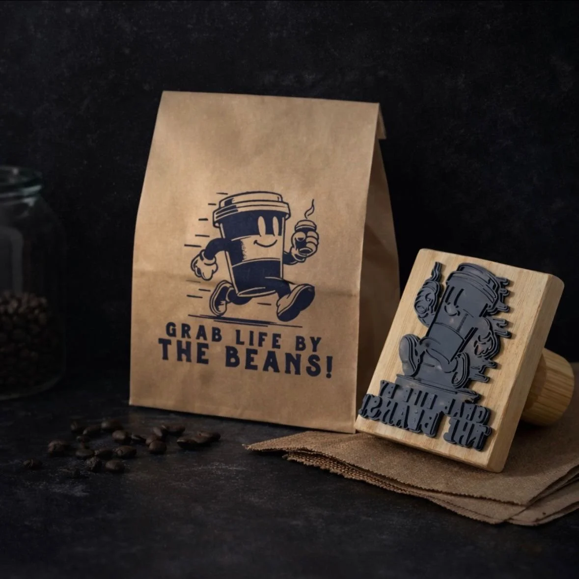 Paquete de papel con diseño de una taza de café y la frase "Grab life by the beans", junto a un sello de goma con el mismo diseño, sobre un paño de tela, al fondo una jarra y en primer plano algunas semillas de café.