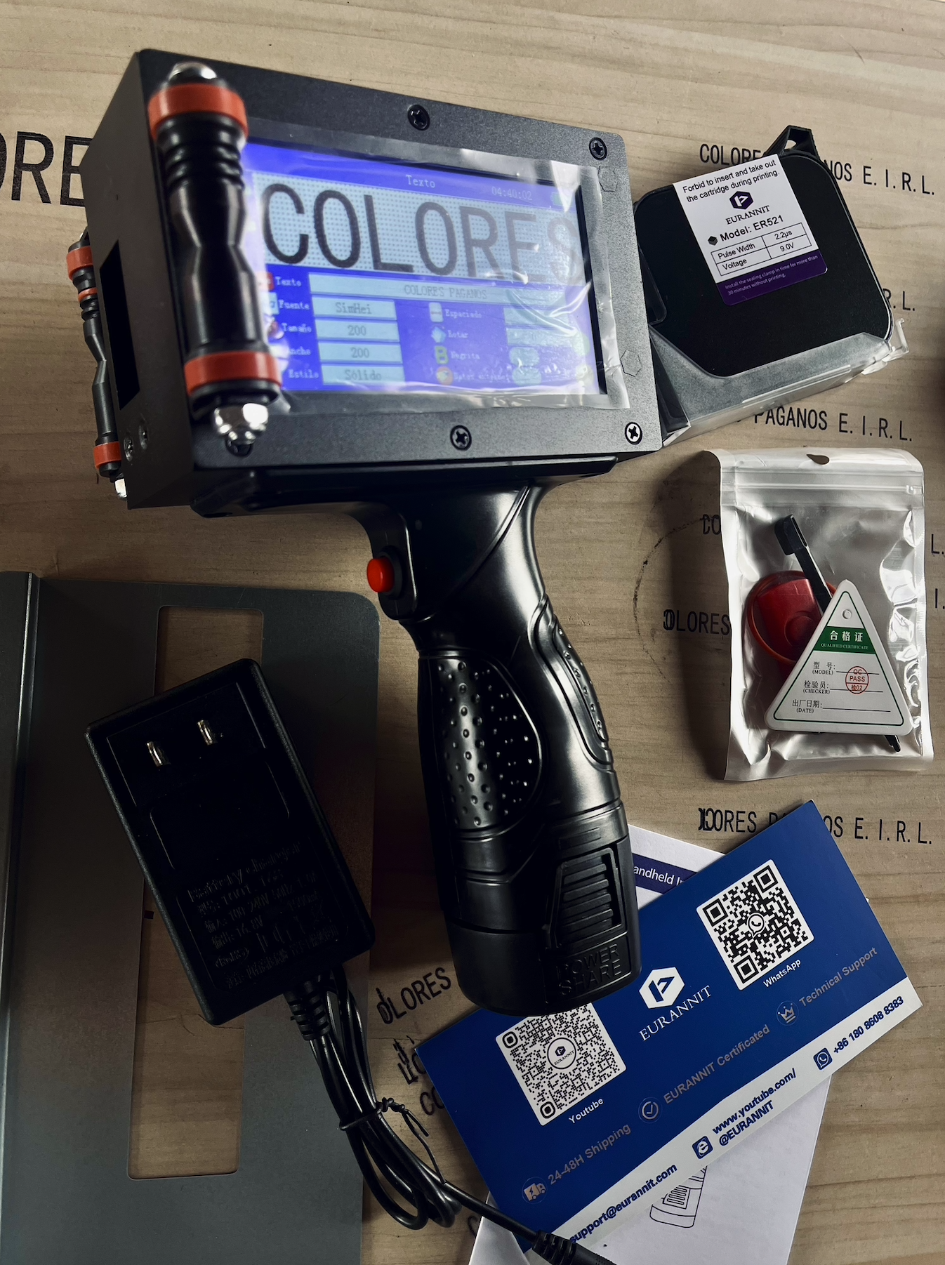 Dispositivo de escáner de códigos de barras con pantalla que muestra la palabra 'COLORES', acompañado de una impresora de etiquetas, una batería, una tarjeta de presentación y otros accesorios en una mesa.