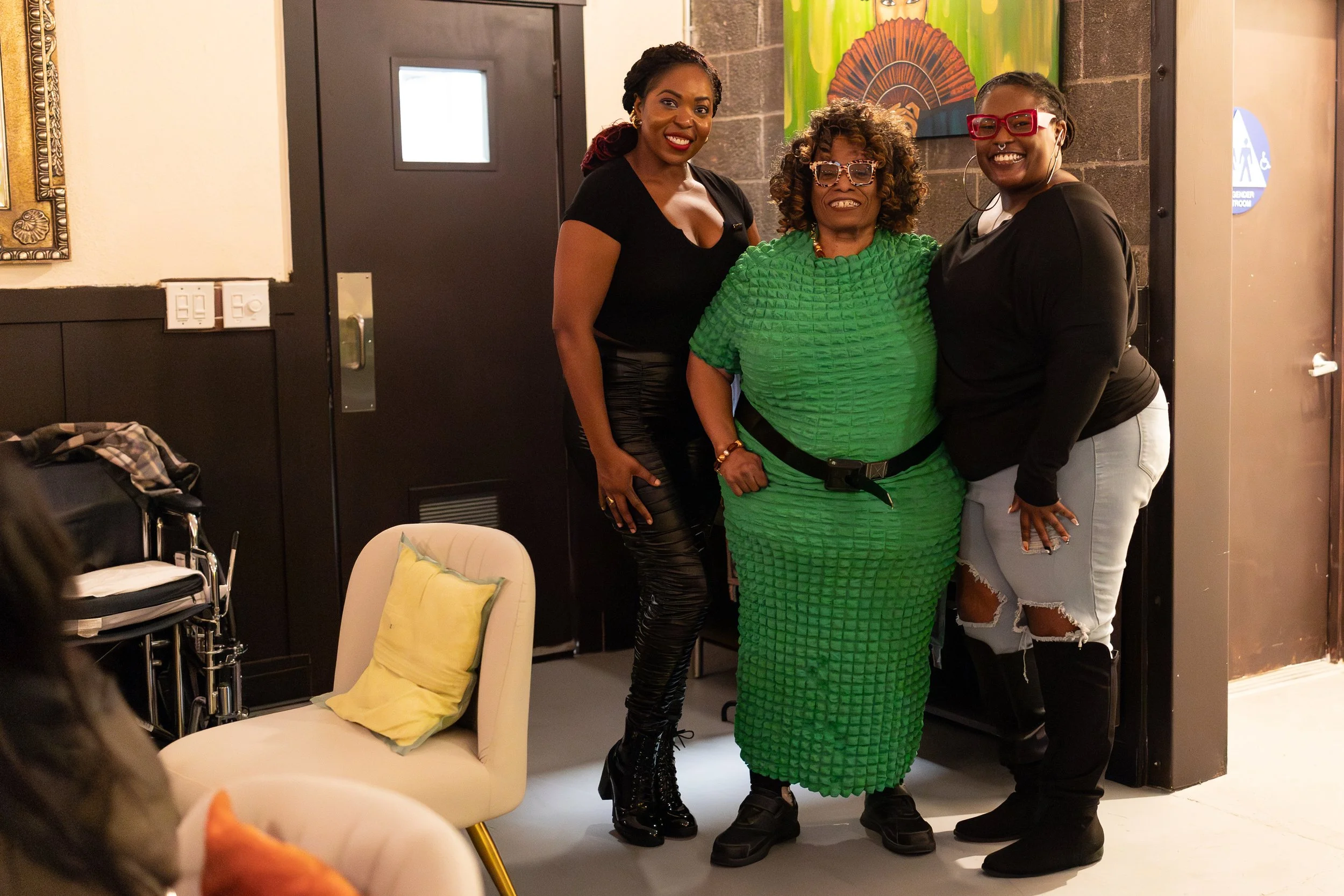 Three people pose together, with one wearing a distinctive textured green dress in the center. 