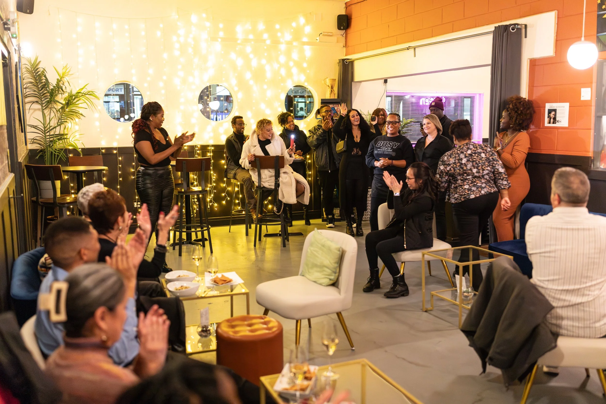 A candid event scene showing people standing and sitting in a lounge space with string lights on the wall creating a warm ambiance. The group appears to be engaged in conversation or presentation, with some people applauding.