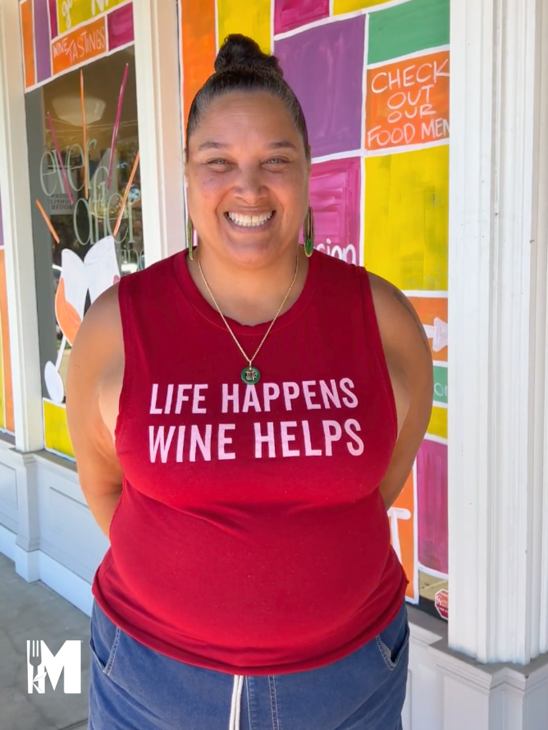 A woman standing in front of a colorful window with in a red shirt with the text "Life happens, wine helps" smiling.