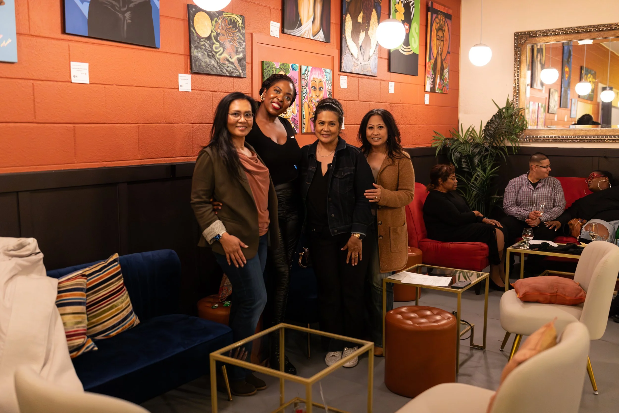 Four people pose together in front of a wall displaying colorful artwork. The space features comfortable seating including a blue velvet couch with striped pillows and modern gold coffee tables.