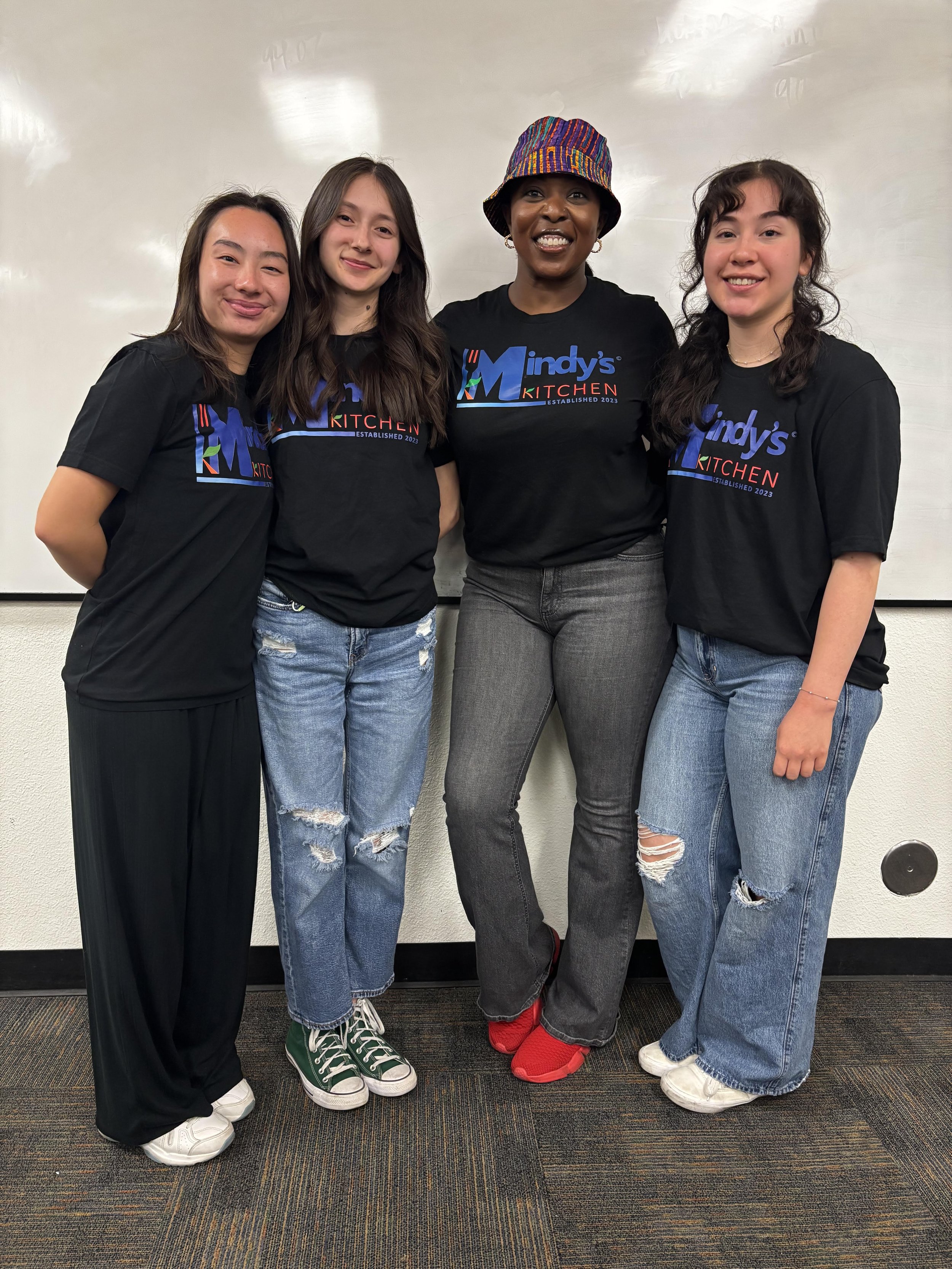Four smiling Mindy’s Kitchen team members pose together in matching branded shirts.