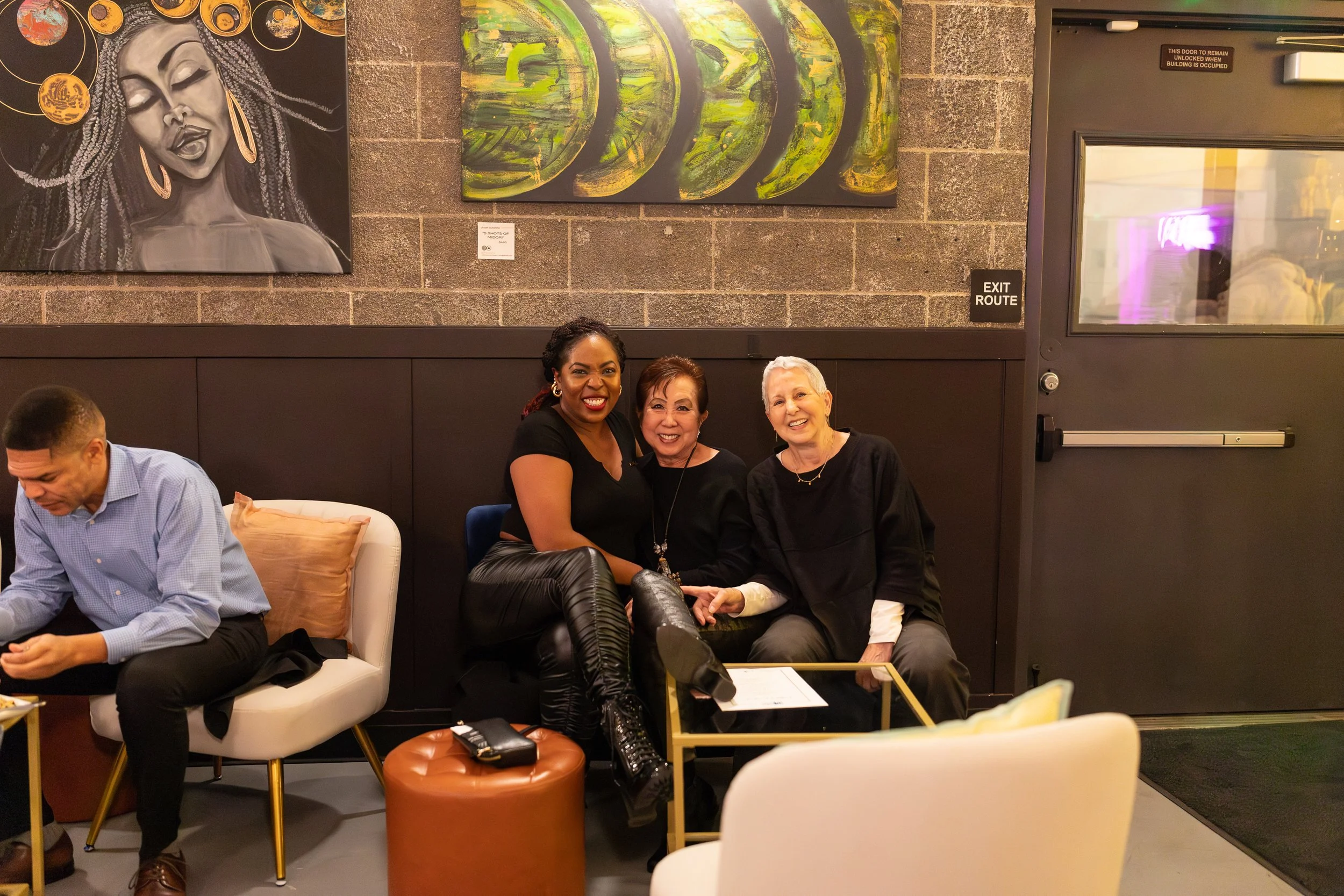 Three people sit together on modern white seating furniture. Behind them is artwork including a dramatic black and white portrait and a series of green circular paintings.