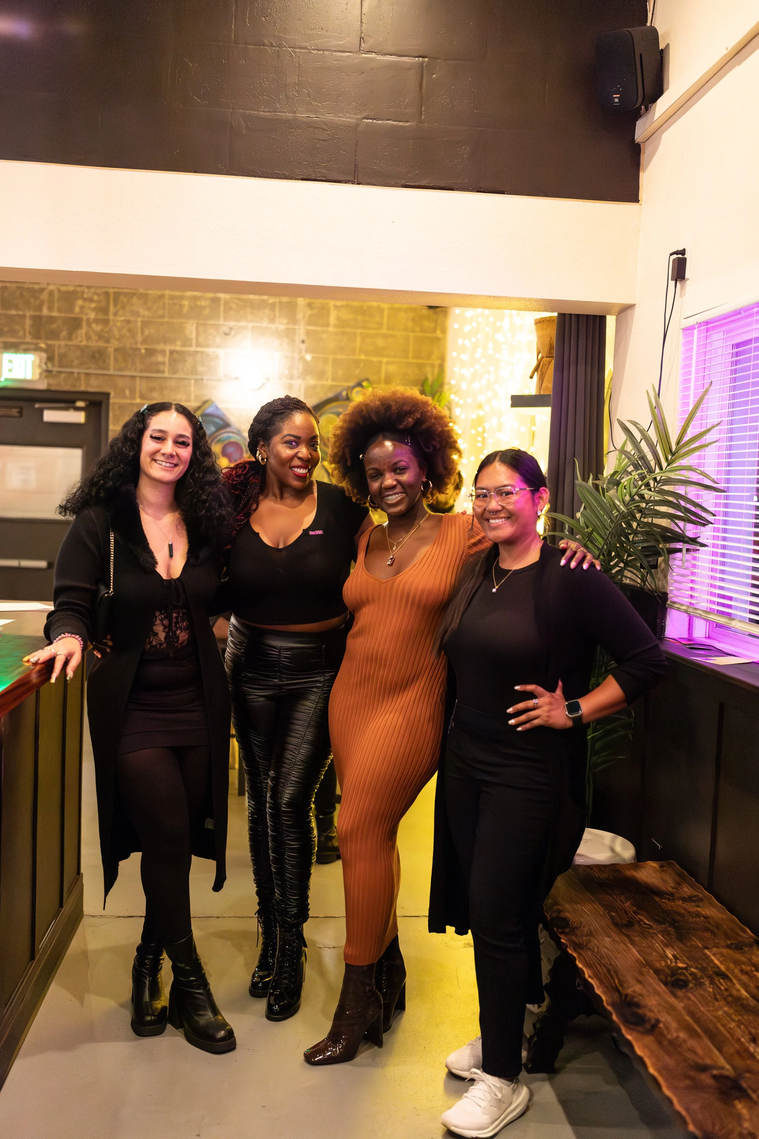 Four women pose together in what appears to be a lounge or bar setting. They're dressed stylishly, with one wearing an orange dress, and the others in black outfits. The space has decorative lighting and a plant in the background.