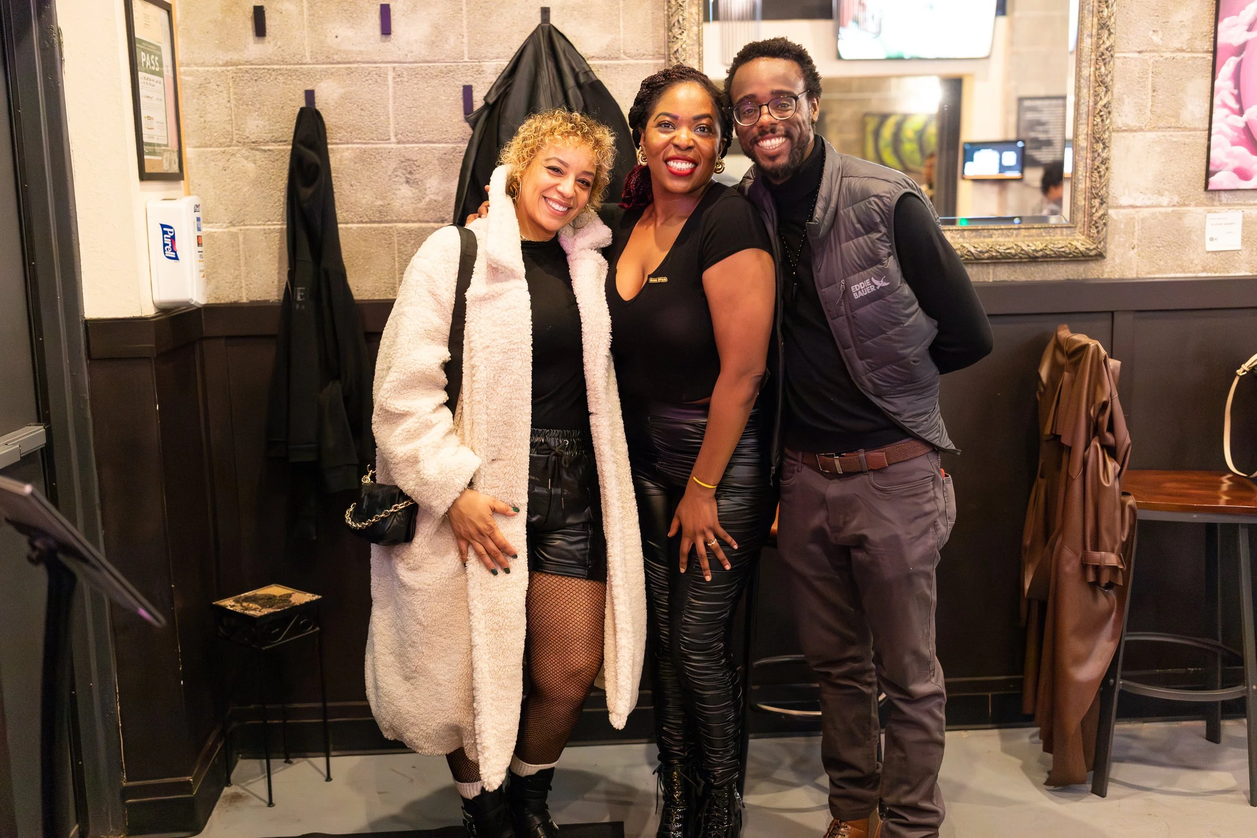 Three people pose together against a grey wall. One person wears a long white fuzzy coat, another wears leather-look pants, and all are smiling warmly at the camera.