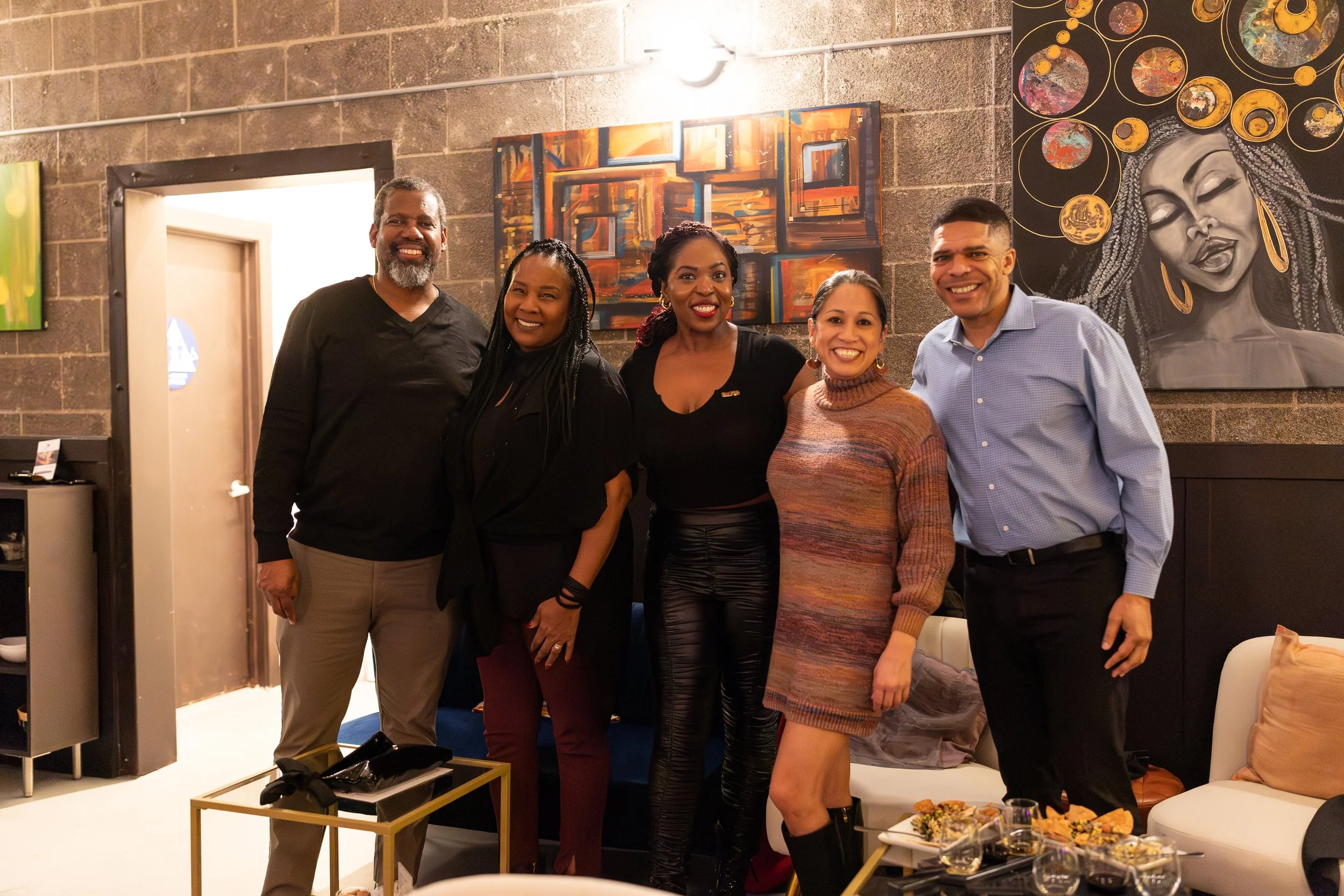 Five people pose together in front of an artistic wall featuring paintings, including a striking portrait of a woman with braids. The space has industrial-style concrete walls and modern decor.
