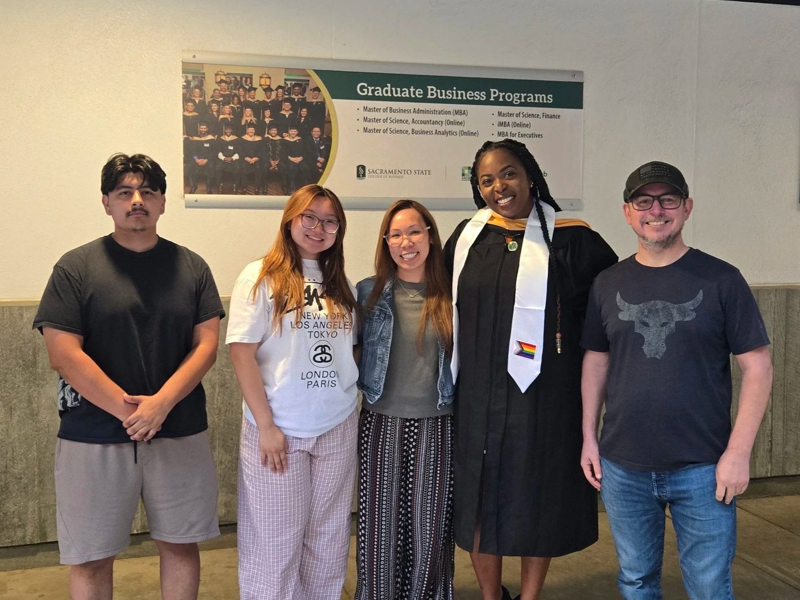 Five people pose together at Sacramento State, including a smiling graduate wearing a cap, gown, and stole.