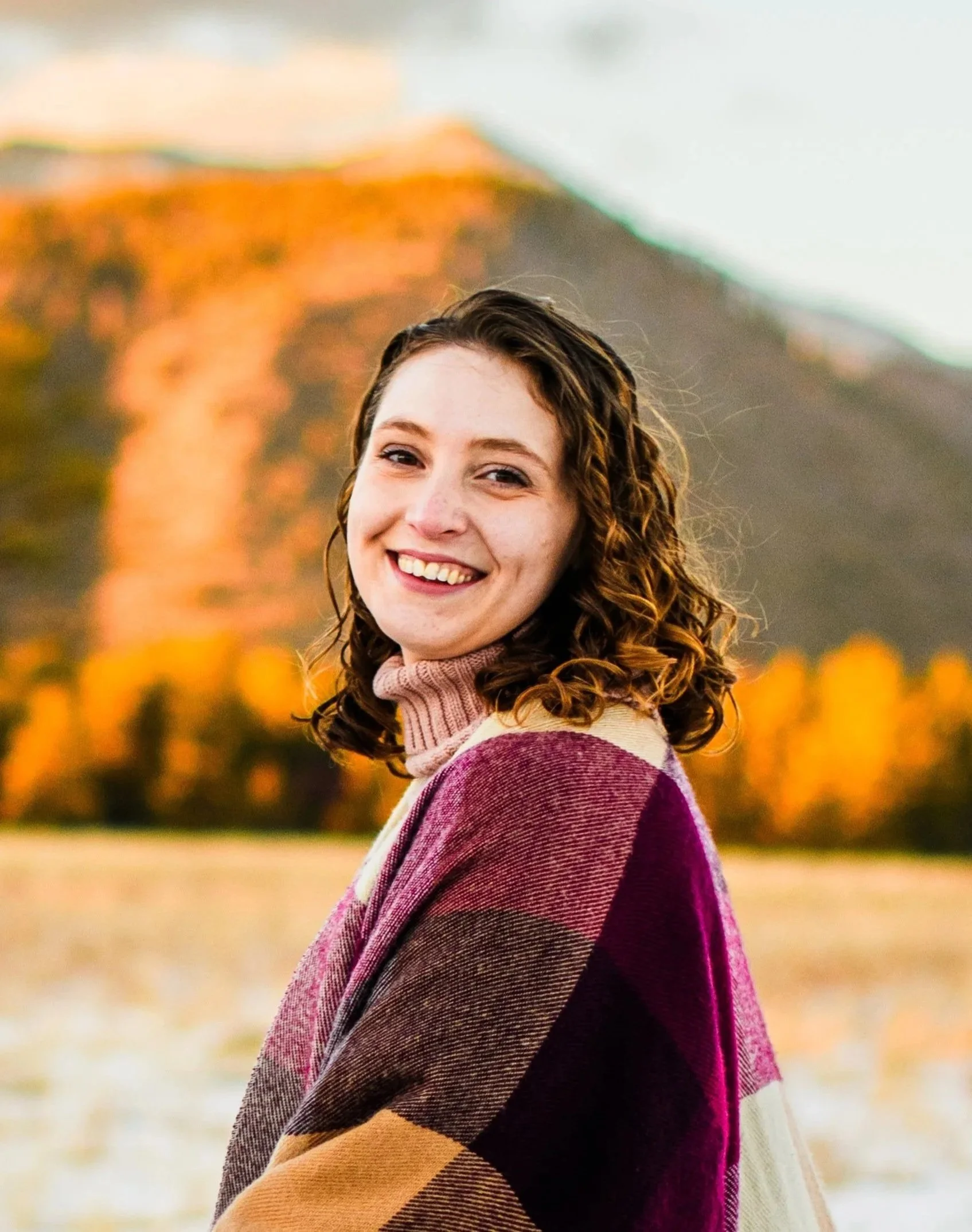A smiling young woman with curly brown hair wearing a multicolored scarf stands outdoors during autumn, with blurred fall foliage and a hill in the background.